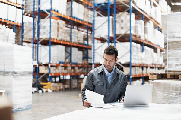 A young man using his laptop and looking over his notes while working in a warehouse